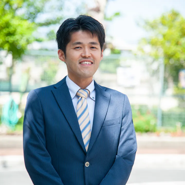 代表:巽 良太の写真。ネイビーのスーツを着て、黄色とネイビーのストライプ柄のネクタイを締め、微笑んでいる。